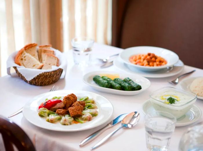 A table adorned with a white tablecloth, featuring an assortment of dips, bread, meats, and stuffed grape leaves, basking in sunlight by a shaded window.