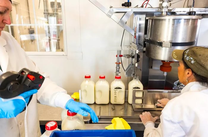 Two women in white coats and blue gloves operate a bottle-filling machine.