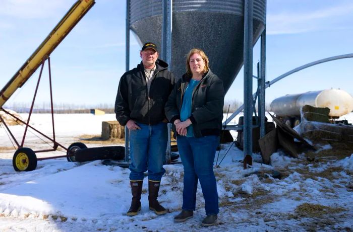 A man in work boots and a black jacket stands next to a woman, also dressed in boots and a jacket, as they pose for the camera in front of industrial equipment on snow-covered ground.