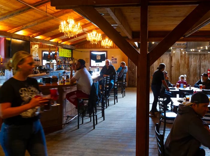 In a warmly lit wooden room adorned with chandeliers, men and women gather at tables and along the bar.