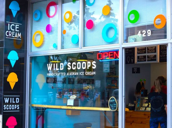 The exterior of this ice cream shop is adorned with vibrant abstract dots, a banner showcasing illustrated ice cream cones, and the name Wild Scoops prominently displayed on the window. A chalkboard menu highlights the daily specials.