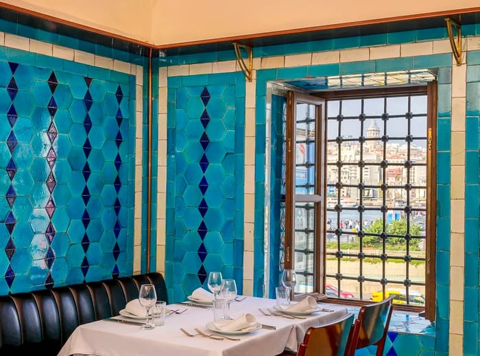 A cozy corner table adorned with dark leather seating, under vibrant blue tiled walls, offering a window view of Istanbul.