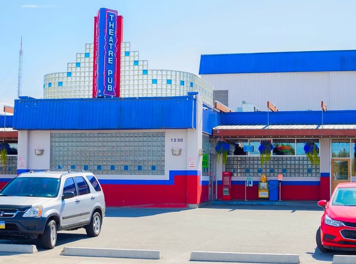 An exterior view of a restaurant and theater featuring glass bricks, vibrant red and blue colors, and a prominent vertical sign proclaiming the “theatrepub.”