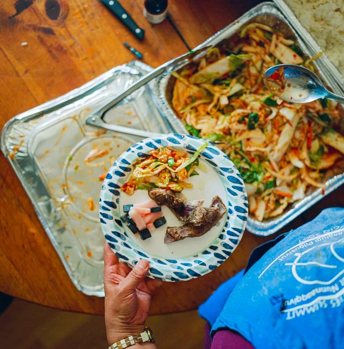 A hand grips a paper plate while holding tongs to serve portions of white-green kimchi, pink maktak, and rice.