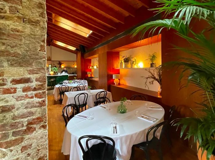 The restaurant features an interior adorned with tables set with white tablecloths beneath a sloped ceiling dotted with skylights. An old brick wall adds character, complemented by soft lighting and wood accents in another wall filled with plants.