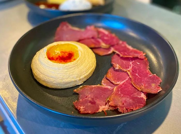 A generous serving of hummus, featuring a well of oil in the center, surrounded by slices of cured meat.