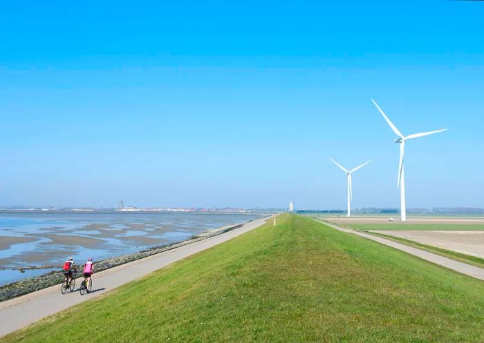 Two cyclists traverse a pathway beside water and wind turbines in Schouwen-Duiveland, Zeeland, the Netherlands.