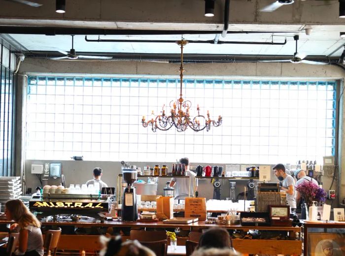 The cafe features a stylish interior highlighted by a large wall of glass bricks behind a lengthy coffee bar, complemented by an elegant chandelier.