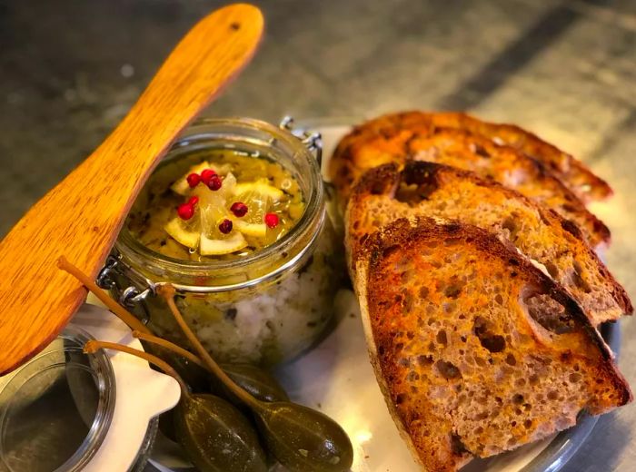 A swing-top jar filled with a chunky spread sits next to slices of toasted bread and pickled vegetables.