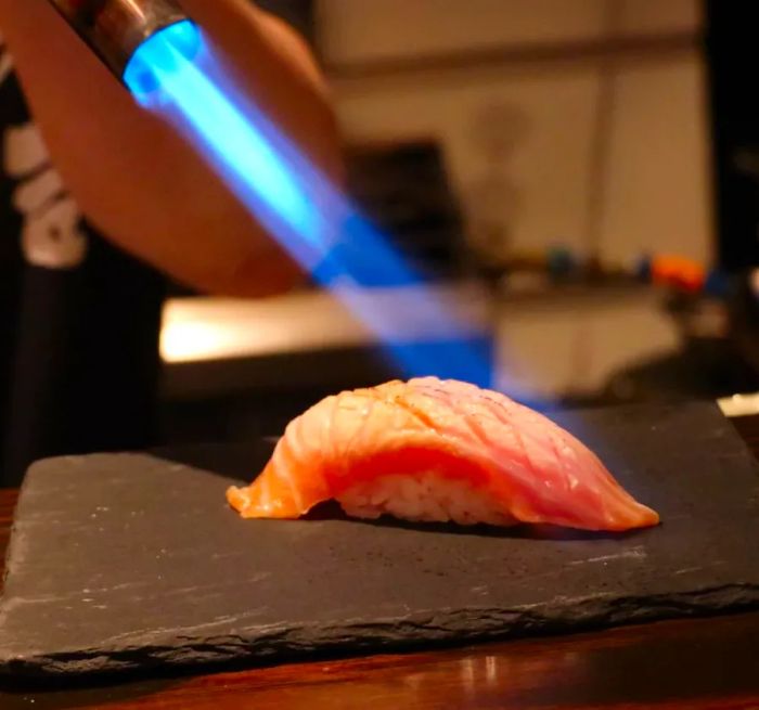 A chef expertly torches a piece of nigiri on a dark countertop.