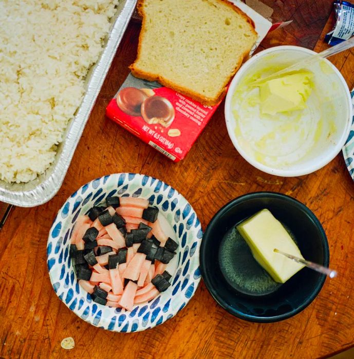 Some bowls are filled with pink and black pieces of maktak, alongside side dishes of butter, a slice of bread, a pan of rice, and a red box of Girl Scout cookies.