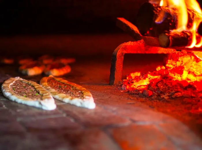 Two pides baking in an oven with a roaring fire beside them.