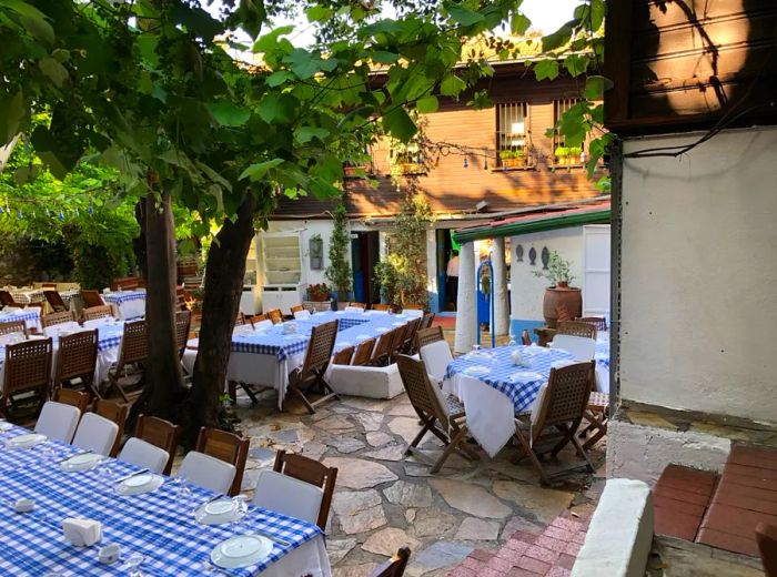 Tables draped in gingham tablecloths are set on a stone patio, surrounded by shading trees, with a grand house encircling the courtyard.