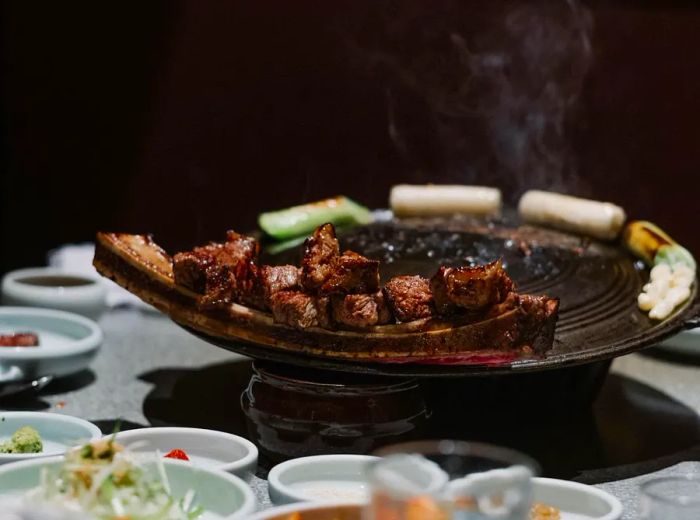 A long beef rib with grilled meat slices resting on the edge of a grill pan, surrounded by banchan.