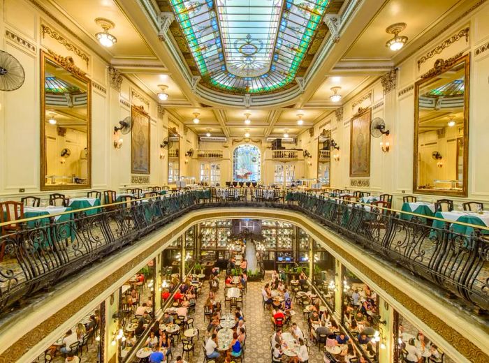 The cafe features an elaborate two-story design with large mirrors adorning the walls and a stained glass skylight, while diners enjoy their meals on the bustling ground floor below.