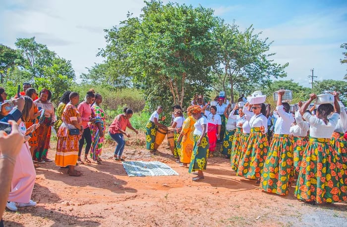 Outside, individuals wearing skirts balance food on their heads, forming two groups facing each other across a cloth.