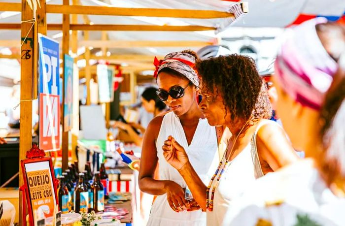 Two individuals explore an outdoor stall in a sunlit market.