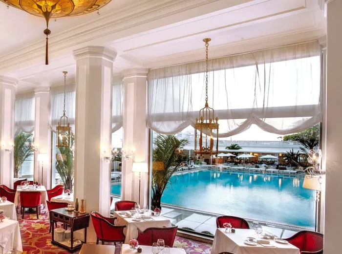 A dining area with a view of the pool, featuring red upholstered armchairs around white tablecloth-covered tables and elegant chandeliers overhead.