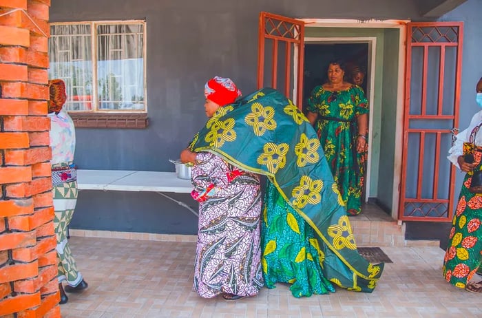 A woman draped in a blanket or shroud walks closely behind another woman.