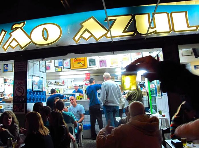 A vibrant night at the bar, where patrons gather outside under the illuminated sign that reads Pavão Azul.