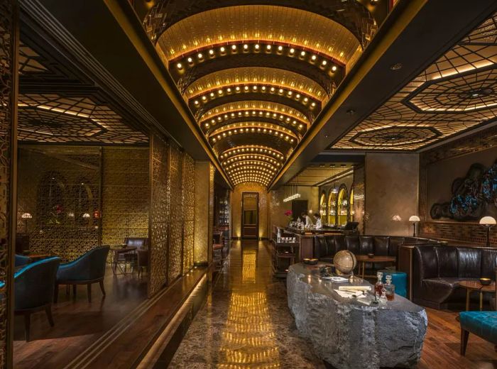 The interior of the bar is dimly lit, featuring gold accents and shimmering lights that follow the curves of an arched ceiling. Black leather booths create an inviting atmosphere, complemented by private dining areas separated by stylish geometric partitions.