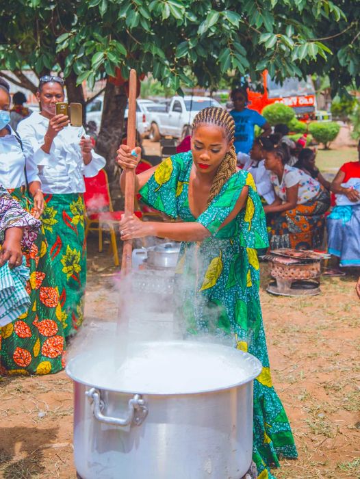 A woman stirs a pot with a long wooden utensil outdoors, surrounded by a group of onlookers.