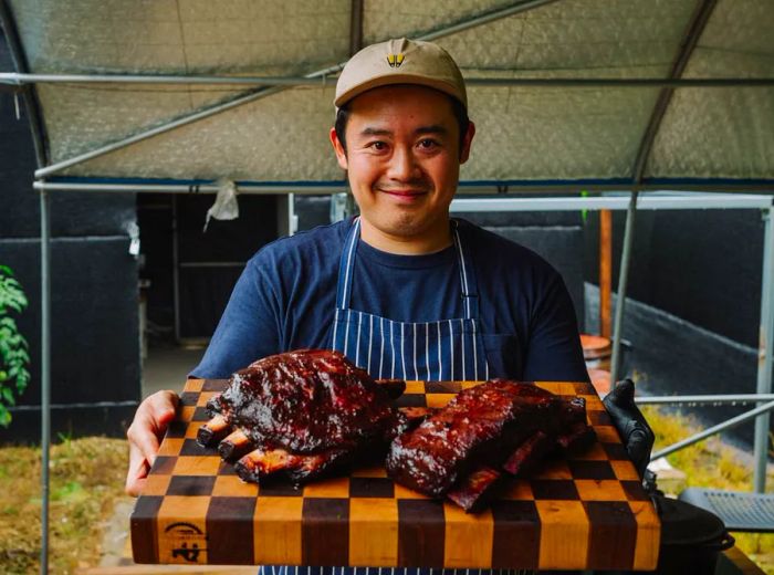 A chef in an apron and baseball cap proudly displays a wooden platter filled with grilled meat.