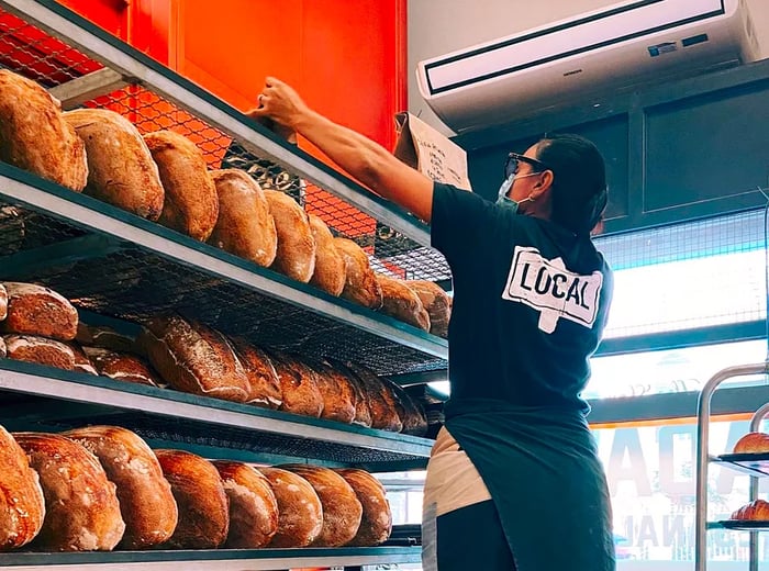 A staff member in a branded shirt uses a ladder to access the top shelf of a large metal shelving unit filled with various loaves of bread.