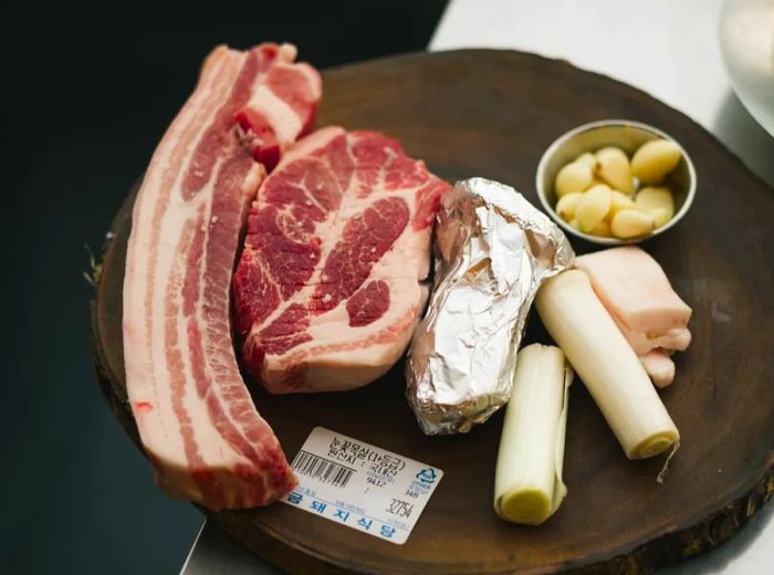 Two generous cuts of meat presented on a wooden platter alongside a foil packet, raw garlic, and assorted vegetables.