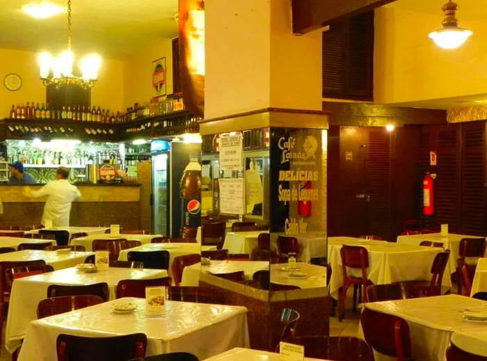 An inviting restaurant space featuring mirrored columns, elegant chandeliers, white table linens, and an open kitchen at the rear.