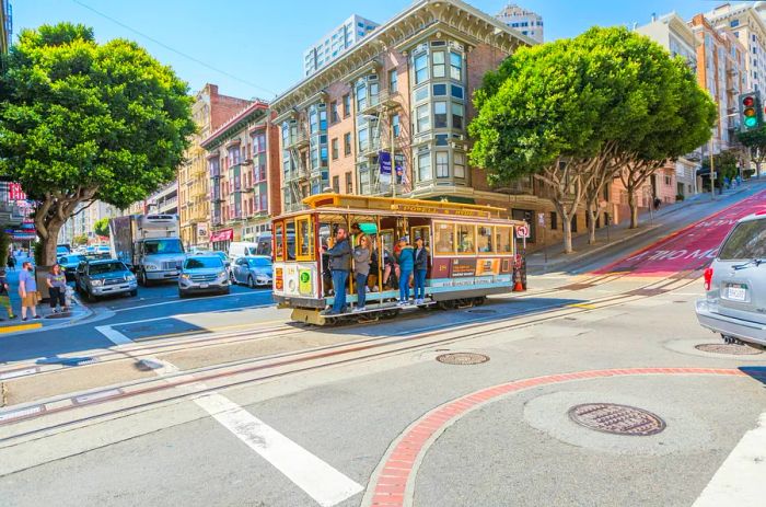 A cable car filled with passengers navigates a bustling intersection on a sunny day.