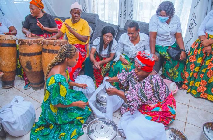 Two women are seated on the floor in front of food containers as drummers perform in the background.