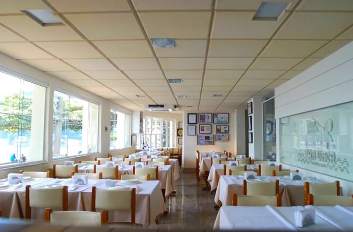 A bright and airy restaurant interior featuring expansive windows along one wall, a paneled ceiling, midcentury-style chairs, and a striking piece of artwork opposite the windows.