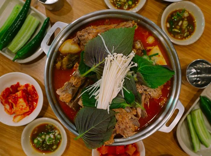 An overhead view of a pot filled with red pork stew, garnished with greens and enoki mushrooms, surrounded by banchan.