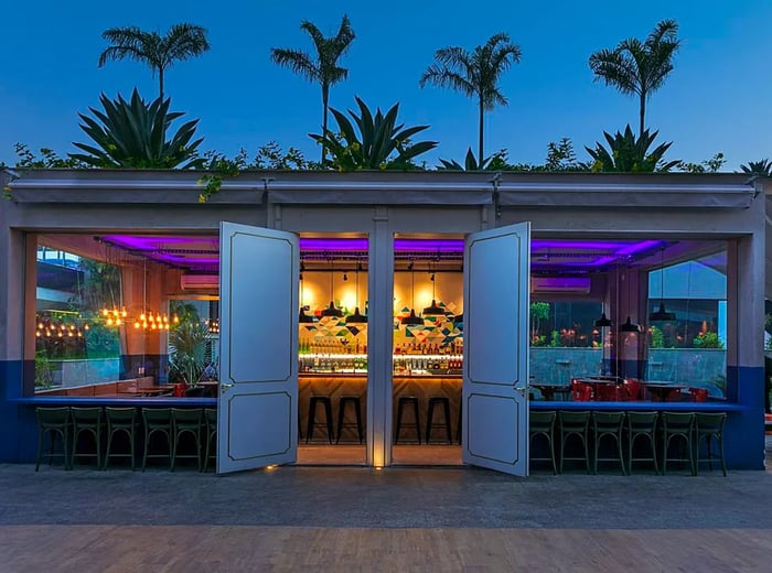 The exterior of a restaurant illuminated at night, with palm trees gently swaying above.