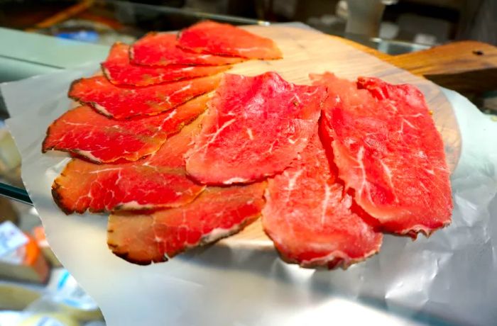 Three images depict: slices of red marbled meat on a wooden serving board, balls of cheese displayed on a labeled shelf, and a midday spread featuring small bites, wine, and decorative gourds.