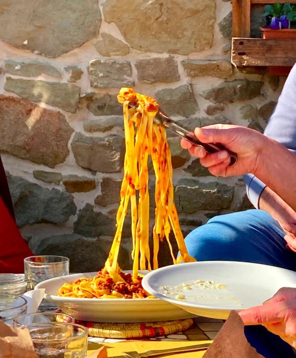 A hand lifts a forkful of pasta from a plate set on a sunlit table.