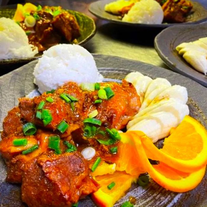 Tender chicken and pork in a savory sauce, garnished with chopped scallions, accompanied by orange wedges and rice.