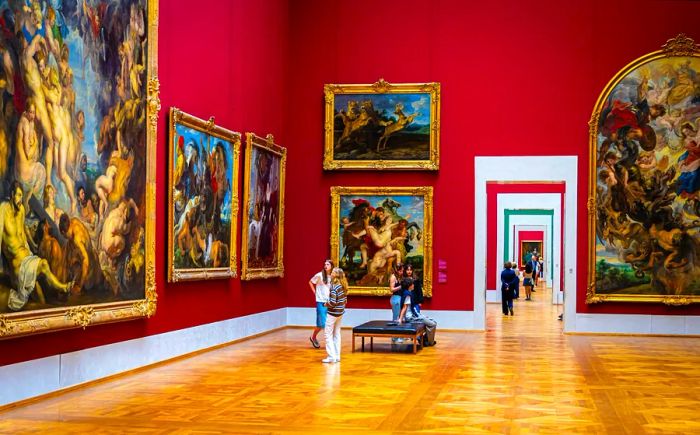 Visitors stroll through galleries featuring red walls adorned with grand old master paintings at the Alte Pinakothek museum in Munich.