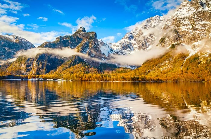 An autumn scene with golden trees and steep, snow-covered peaks shrouded in clouds beside the Königssee, Bavaria