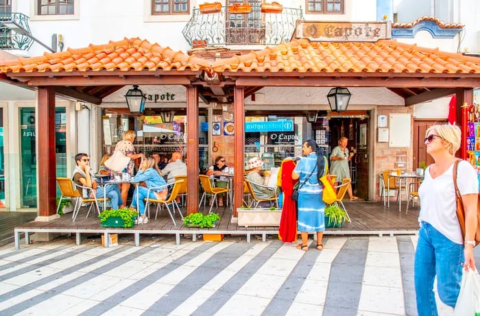 A pedestrian walks past a sunlit restaurant with diners enjoying meals at outdoor tables shaded by shingled awnings.