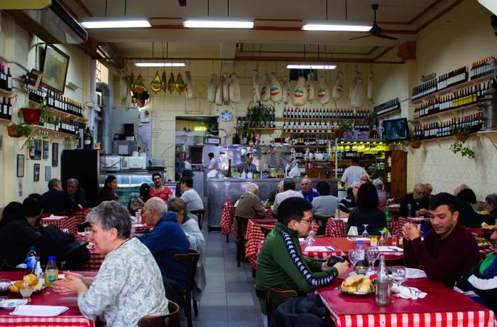 An interior shot of a bustling restaurant filled with patrons at red checkered tablecloths, large pieces of meat hanging from the ceiling, cooks working behind a counter in the background, and shelves lined with bottles of wine on the walls.