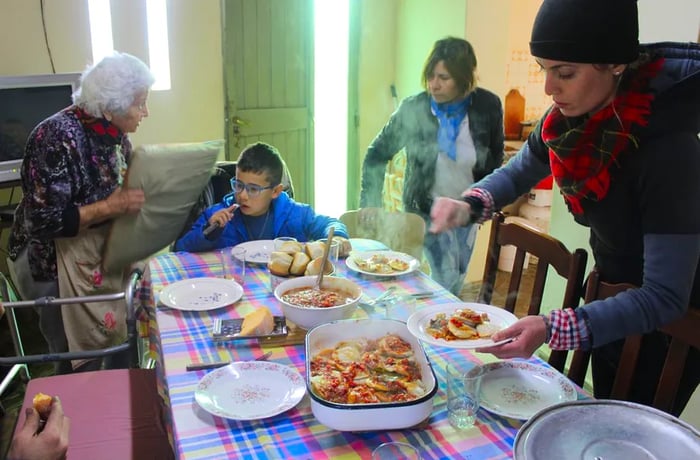 A family gathers around a table laden with dishes, including a steaming, sauce-covered baked dish releasing fragrant waves of steam.