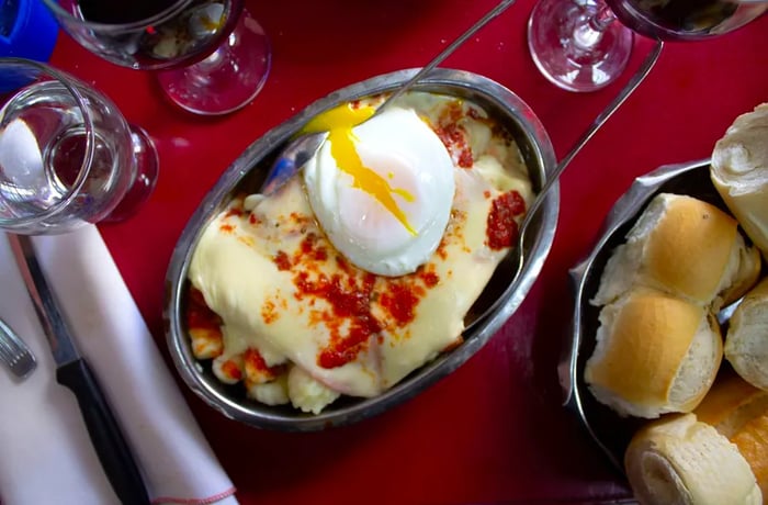 From above, a metal plate with tongs, hiding the ñoquis beneath a layer of cheese and a runny egg.