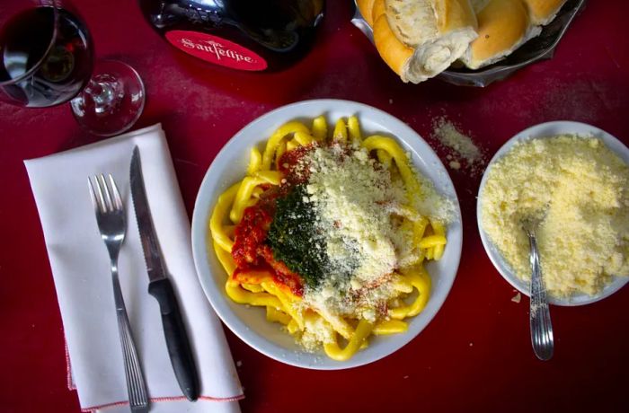 An overhead view of a pasta dish generously topped with grated cheese and sauces, alongside a bowl of additional grated cheese.