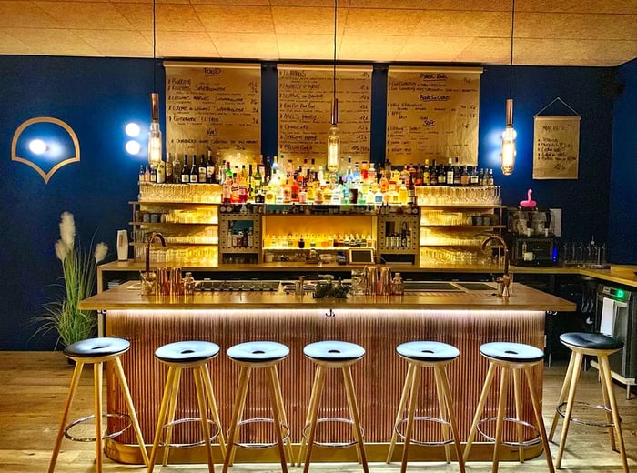 Stools line a chic, illuminated bar against a deep blue wall, with menus displayed on butcher paper rollers attached to the back wall, illuminated bottles on the bar, and elegant gold fixtures atop a wooden bar surface.