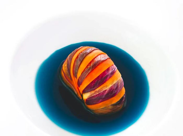 A vivid lobster tail rests in a deep, dark blue sauce within a nearly invisible white soup bowl against a neutral backdrop.