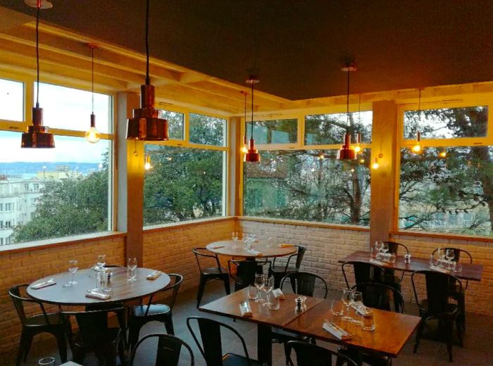 The dining room is empty, featuring wooden tables of various shapes, low brick walls beneath the windows, and softly lit by pendant lights, with trees and a cityscape visible outside.