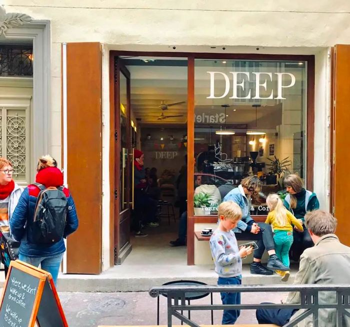 The cafe facade features the name DEEP prominently displayed in large letters above the window, with patrons mingling both outside and inside.