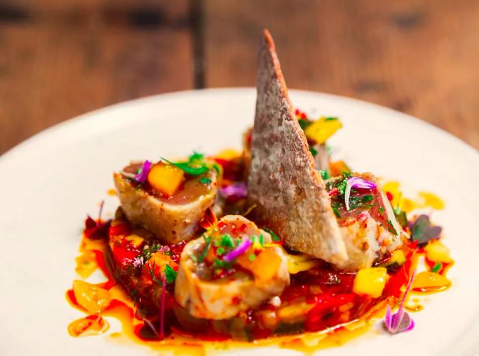 A plate features an arrangement of cooked fish slices encircling a prominent piece of fish skin, reminiscent of a shark fin, rising from the center. The dish is adorned with a vibrant red sauce and decorative garnishes on a pristine white plate.